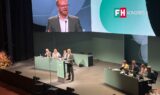 En mand i jakkesæt holder tale fra talerstolen på en FH-konference, hvor deltagerne sidder på scenen, og en stor skærm viser taleren bag ham.