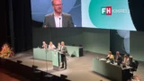 En mand i jakkesæt holder tale fra talerstolen på en FH-konference, hvor deltagerne sidder på scenen, og en stor skærm viser taleren bag ham.