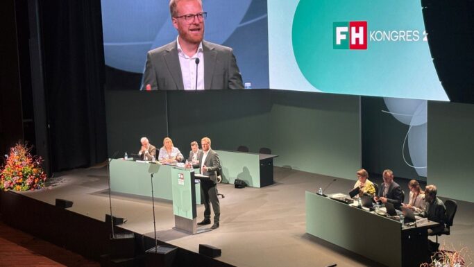 En mand i jakkesæt holder tale fra talerstolen på en FH-konference, hvor deltagerne sidder på scenen, og en stor skærm viser taleren bag ham.