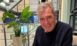 En smilende ældre mand ved navn Niels sidder indenfor ved siden af en potteplante og et skilt for Re:Cognition Health, et ekspertcenter for demenspleje.