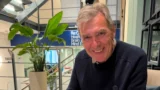 En smilende ældre mand ved navn Niels sidder indenfor ved siden af en potteplante og et skilt for Re:Cognition Health, et ekspertcenter for demenspleje.