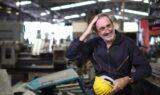 En smilende mand i en mørk overalls og briller står med en gul sikkerhedshjelm i hånden, mens han holder en pause på en fabrik og tænker over kravene til alderspension.