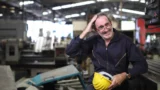 En smilende mand i en mørk overalls og briller står med en gul sikkerhedshjelm i hånden, mens han holder en pause på en fabrik og tænker over kravene til alderspension.