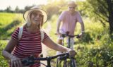 Et ældre par med hatte og stribede skjorter, der smiler, mens de går med deres cykler på en solbeskinnet sti, og tænker over pensionsplanlægning og pensionsskat.