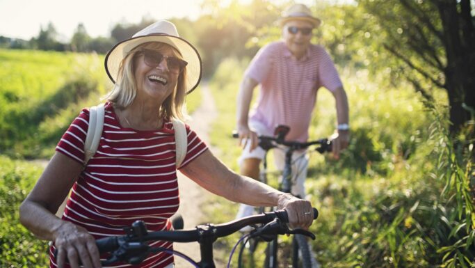 Et ældre par med hatte og stribede skjorter, der smiler, mens de går med deres cykler på en solbeskinnet sti, og tænker over pensionsplanlægning og pensionsskat.
