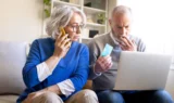 En ældre kvinde taler i sin smartphone, mens en ældre mand kigger på skærmen på en bærbar computer og holder et kreditkort i hånden, hvilket illustrerer risici som svindel.