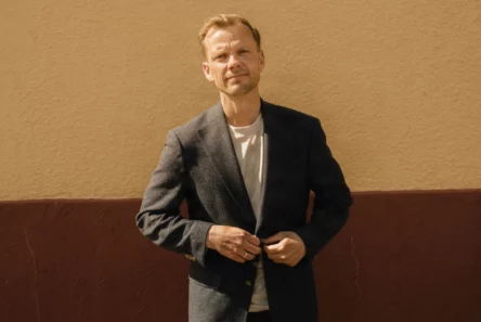 En mand i blazer og hvid T-shirt knapper sin jakke, mens han står op ad en tofarvet væg og reflekterer over den aktuelle danske økonomi.
