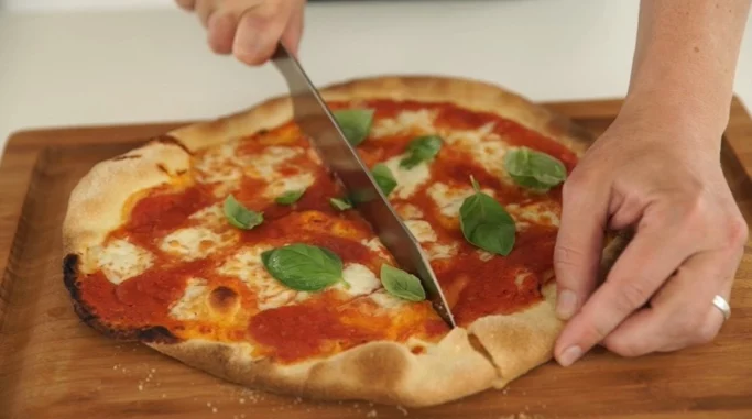 Hænder, der bruger en kniv til at skære en ægte Pizza Margherita med frisk basilikum og mozzarella på et træskærebræt.