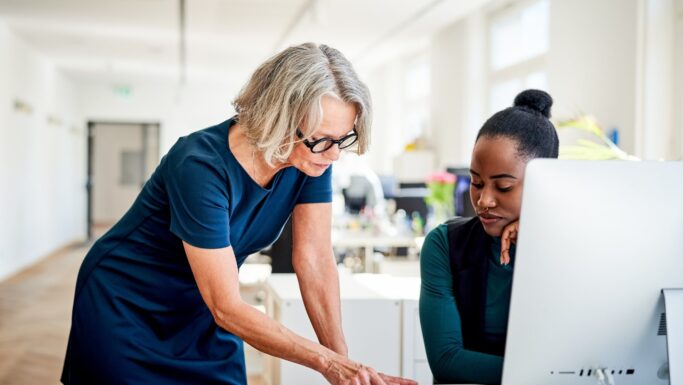 En kvinde i en blå kjole læner sig frem over et skrivebord for at rådføre sig med en erfaren kollega, der sidder ved en computer, hvilket understreger et aktivt engagement på arbejdspladsen.