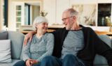 Et ældre ægtepar, der nyder deres pension, sidder på en sofa, smiler og ser på hinanden.
