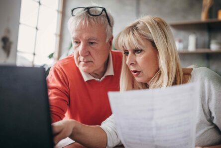Et ældre ægtepar kigger på et dokument, mens de sammen arbejder på en bærbar computer for at gennemgå regeringens spareplan.