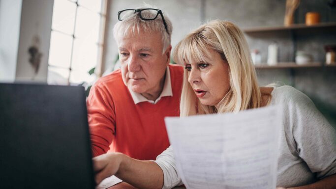 Et ældre ægtepar kigger på et dokument, mens de sammen arbejder på en bærbar computer for at gennemgå regeringens spareplan.