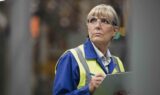 En kvinde iført sikkerhedsbriller og en refleksvest gennemgår de nye regler om alderspension, mens hun tager noter på et skriveunderlag i et industrimiljø.