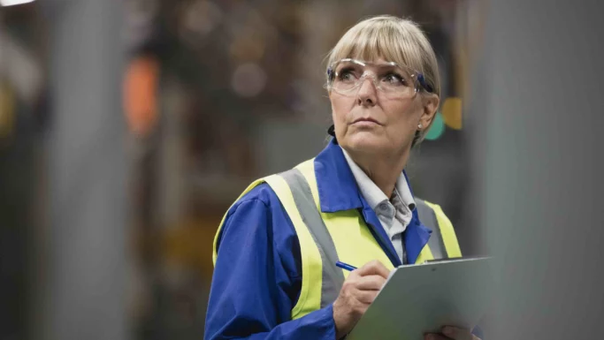 En kvinde iført sikkerhedsbriller og en refleksvest gennemgår de nye regler om alderspension, mens hun tager noter på et skriveunderlag i et industrimiljø.
