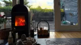 En hyggelig stue med en tændt pejs, et puslespilbord, stearinlys og sten. Udenfor er der en træterrasse med havemøbler og grønne planter – perfekt for dem, der ønsker at sælge eller købe sommerhuse på trods af de høje priser.