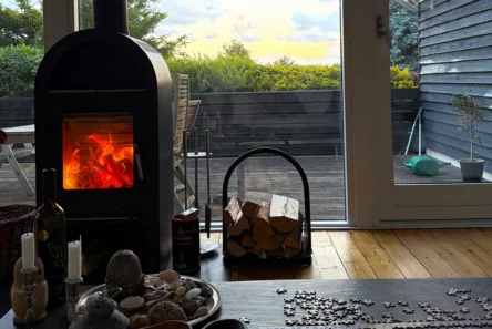En hyggelig stue med en tændt pejs, et puslespilbord, stearinlys og sten. Udenfor er der en træterrasse med havemøbler og grønne planter – perfekt for dem, der ønsker at sælge eller købe sommerhuse på trods af de høje priser.