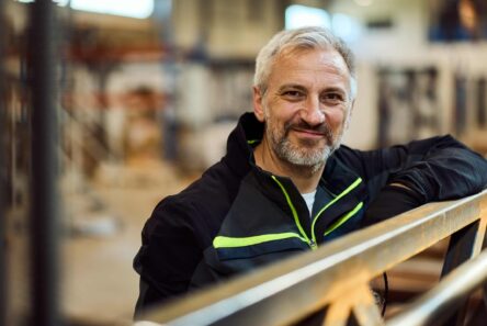 En smilende mand i midten af livet med skæg, iført en sort jakke med neongrønne detaljer, læner sig op ad en værkstedsbygning, mens han overvejer sine muligheder i lyset af de nye pensionsregler.